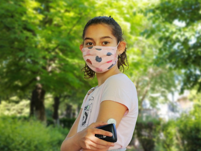 Corona virus and diabetes. Girl with the mask on face checks glucose level in blood with device for continuous glucose monitoring – CGM. She is reading glucose level from the white sensor on left arm.