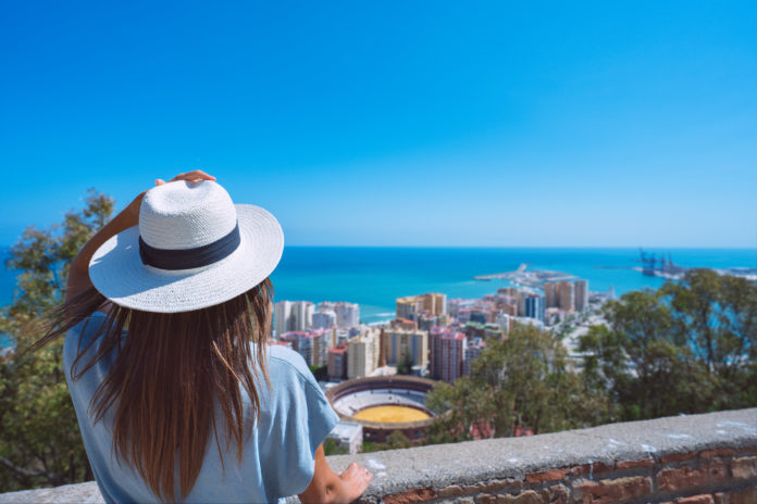 Rear view of young woman traveller with white hat standing in Gibralfaro Castle viewpoint and looking at Malaga cityscape. Summer holiday vacation in Spain